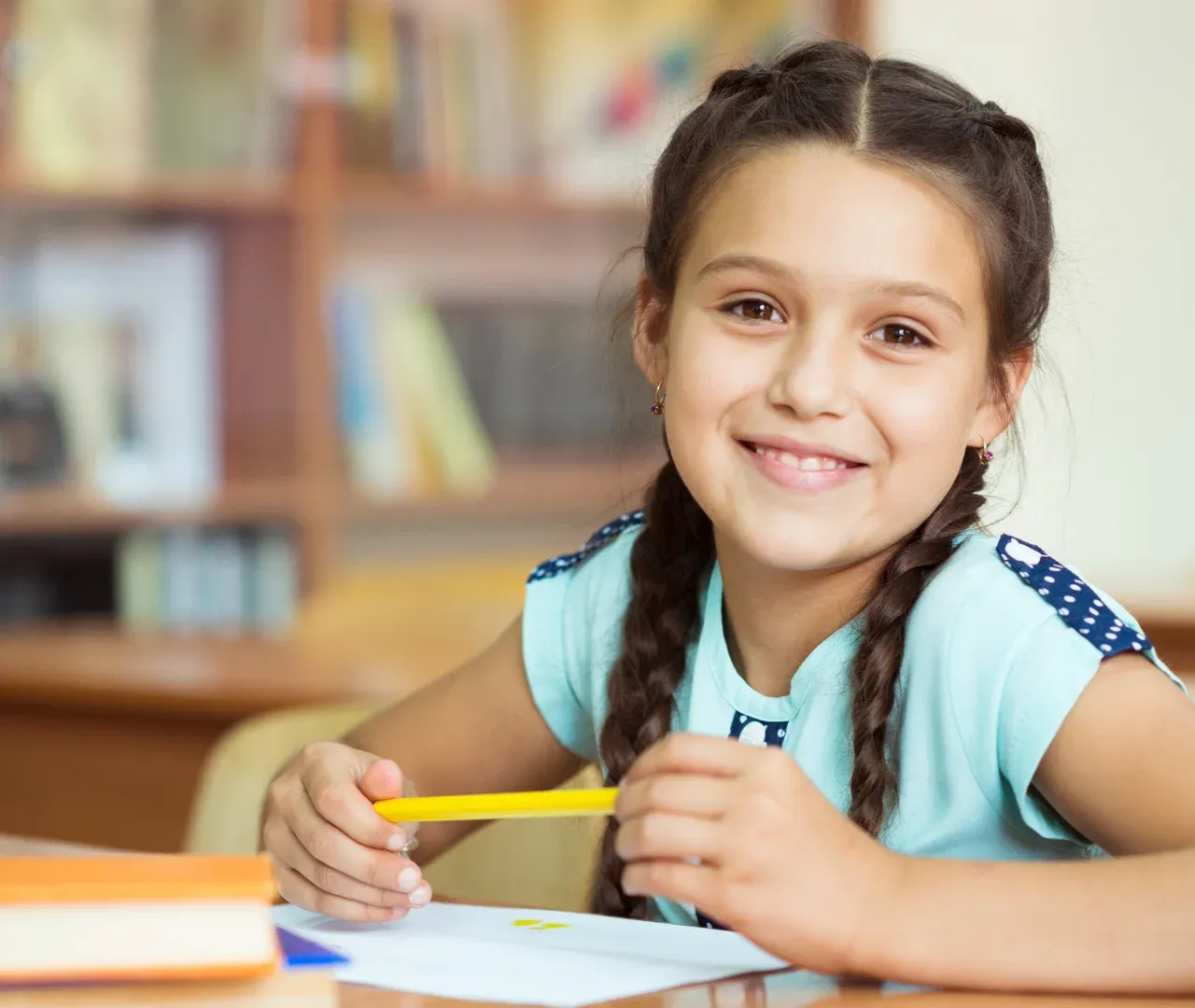A smiling girl with braids holds a pencil at her desk in a classroom